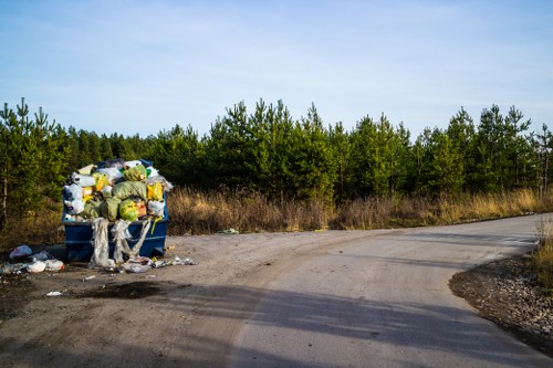A large blue communal rubbish bin situated on the side of a gravel driveway in a rural area, filled with tightly packed plastic bags containing household waste, with some debris spilling onto the ground nearby. The bin is leaning slightly to one side, and the waste inside includes visible plastic, paper, and foam packaging. Surrounding the bin, the ground is uneven with patches of dirt and small stones, and the background features dense green hedgerows and a partly cloudy sky. This scene reflects the type of waste collection service provided by Commercial Waste Palmers Green in the local area, highlighting waste disposal in an outside environment adjacent to a natural landscape.