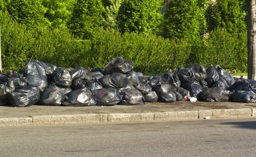 A long row of black garbage bags filled with waste is piled along the edge of a paved road, with some bags leaning against each other. The bags appear to be made of plastic, and their surfaces are slightly shiny, reflecting sunlight. Behind the waste bags, there is dense greenery consisting of tall trees and lush shrubs, indicating a natural or semi-rural environment possibly near Palmers Green. The road in the foreground is grey asphalt with a concrete curb separating it from the waste. The overall scene suggests a rubbish removal situation where Commercial Waste Palmers Green may be involved in clearing waste from outdoor areas or streets in the local area, with the waste bags positioned in a tidy, organized manner awaiting disposal or collection. The lighting is natural daylight, and the waste is situated in an outdoor setting, emphasizing the importance of professional rubbish removal services in maintaining cleanliness in residential or commercial surroundings in the borough.