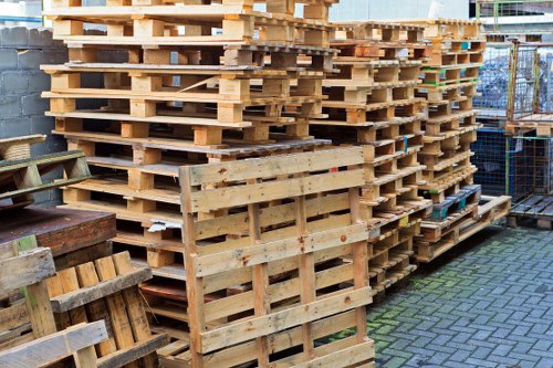 A large stack of untreated wooden pallets arranged outdoors on a paved surface, with some pallets leaning against others and a few smaller pallets placed in front. The pallets are light beige with visible wood grain, and some show darker marks or slight weathering. They occupy the foreground and extend into the background, set next to a grey brick wall, while a metal fence and part of a building are visible behind the pallets. The scene appears to be part of a commercial waste collection site in Palmers Green, with the pallets organized in a tidy manner for eventual disposal or recycling, representative of a typical rubbish removal setting handled by Commercial Waste Palmers Green.
