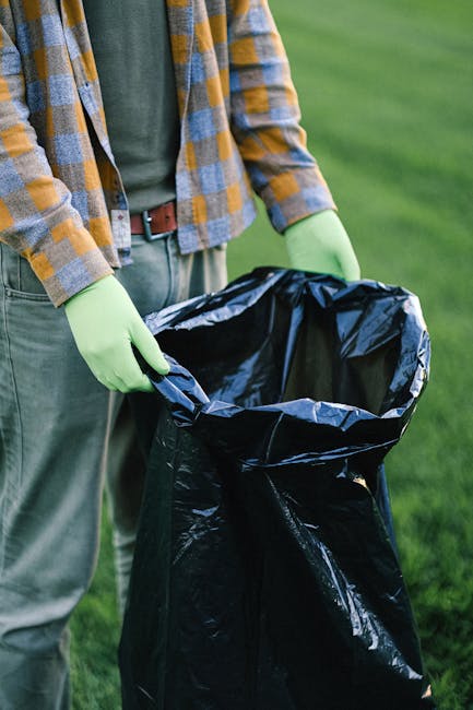 A waste collection worker wearing a high-visibility yellow and orange jacket is standing at the rear of a large red municipal rubbish truck, which is parked on the side of a street. The worker is enga