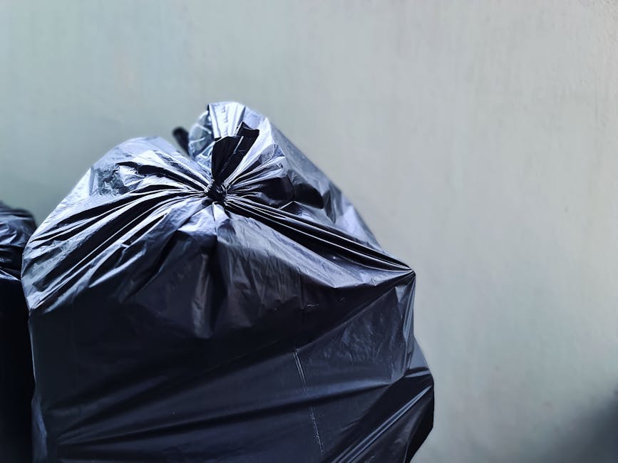 A person dressed in a white protective suit with orange accents, wearing a hood and safety goggles, stands amidst a large outdoor rubbish dump covered with white and multi-coloured waste bags, cardboa