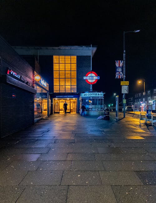 A nighttime street scene in front of Kew Gardens station, with illuminated glass panels behind the station entrance casting a warm glow. The entrance features sliding glass doors, with two individuals